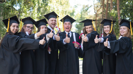 A group of graduates in robes give a thumbs up outdoors. Elderly student.