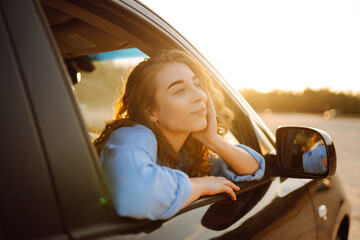 Smiling woman from the window of the car enjoys nature, you feel freedom while traveling by car. Car travel, sunset light. Active lifestyle.