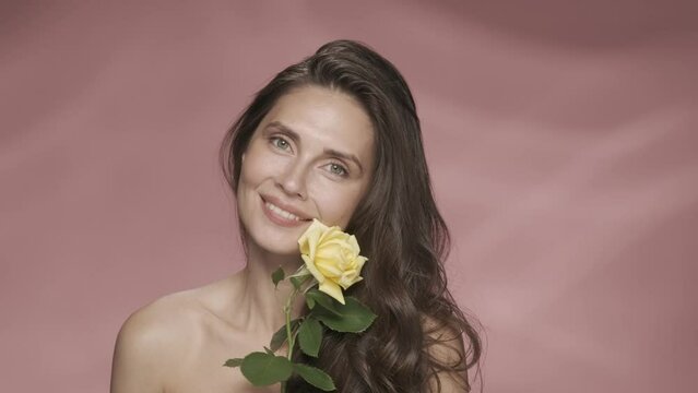 A seminude woman holds a yellow rose on a long stem and inhales its fragrance. Smiling woman holds a rosebud across her face in a studio on a pink background. Spring and summer inspiration. HDR BT2020