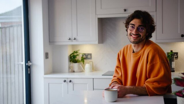 Portrait of smiling young man wearing glasses taking a break with cup of coffee sitting at kitchen counter at home with copy space on right side of frame - shot in slow motion