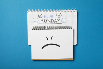 Notepads with text Blue Monday and sad emoji on blue background, top view