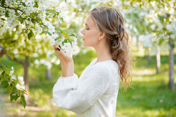 Portrait of a young woman in a white dress with a branch of flowers in her hands against the backdrop of blooming apple trees. The girl poses against the background of flowers in the spring park.