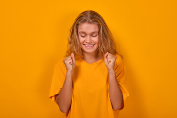 Young overjoyed Caucasian woman millennial makes joyful wave of hands and closes eyes after receiving news of salary increase or new job offer stands posing on plain orange background.