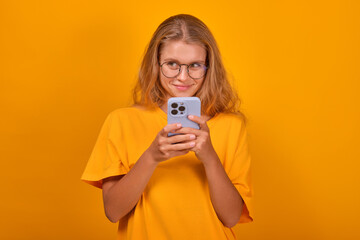 Young cute shy Caucasian woman with phone in hands smiles sweetly and looks away getting to know guys on social networks or dating apps dressed in casual clothes stands in orange studio.