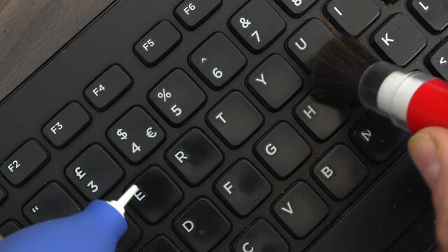 Closeup overhead shot of hands using a blower and brush to clean the keys of a dirty and dusty computer keyboard.