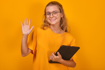 Young happy attractive Caucasian woman teenager waving and saying hello to friends or website visitors and holding tablet computer stands on isolated orange background. Schoolgirl, student