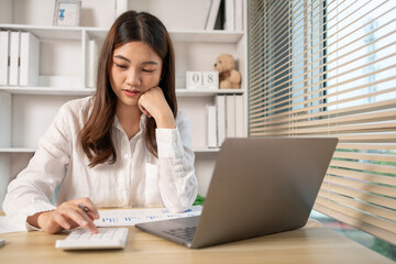 Asian woman working at home, Press the calculator to calculate the income - expenses of the house and use the laptop to record the information and check the accuracy, Home lifestyle.