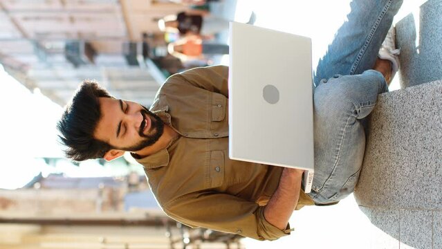 Indian man freelancer working online distant job with laptop sits in city street browsing website chatting outdoors during break. Guy tourist looking at notebook screen sends messages, watching movies