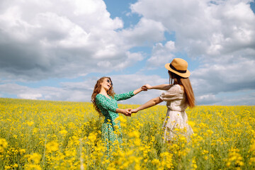 Fototapeta premium Two cheerful girlfriends dressed in stylish dresses are walking through a blooming rapeseed field. The concept of walking, active lifestyle, holidays.
