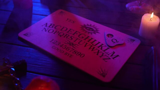 Close up video of a wooden ouija board on a table surrounded by candles, candlesticks and other elements in a muffed light.