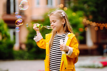 Cheerful little girl in a yellow raincoat blows soap bubbles, plays on a walk in the park in...