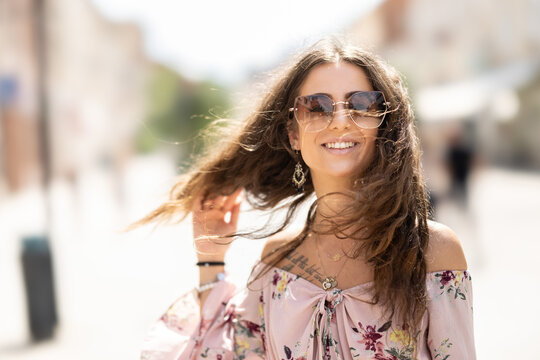 Happy young smiling woman in summer outfit with sunglasses walking in the city with wind blowing her hair