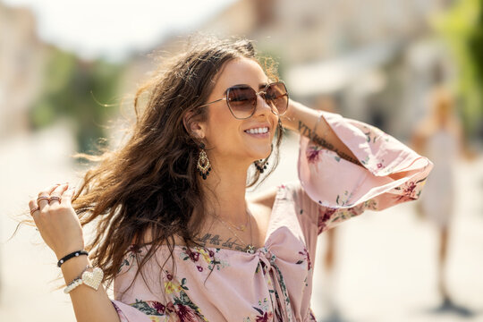 Happy young smiling woman in summer outfit with sunglasses walking in the city with wind blowing her hair