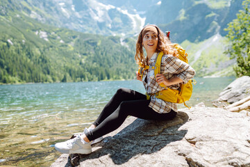 Happy tourist woman with a big yellow backpack enjoys the view of the mountain lake in sunny weather. The concept of hiking, travel, vacation. Active lifestyle.