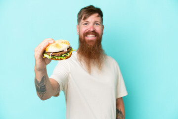 Redhead man with long beard holding a burger isolated on blue background with happy expression