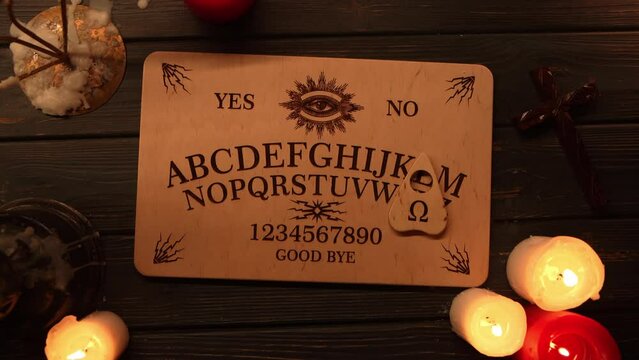 Close up video of a wooden ouija board on a table surrounded by candles and a cross in a muffed light. The planchette is moving on the board.