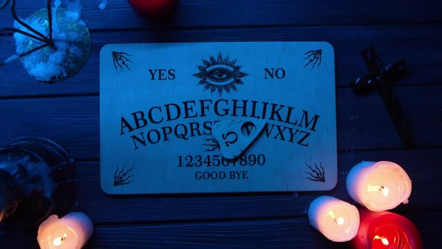 Close up video of a wooden ouija board on a table surrounded by candles and a cross in flashing lights. The planchette is moving on the board.