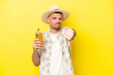 Fototapeten Young caucasian man holding a cocktail isolated on yellow background showing thumb down with negative expression © luismolinero