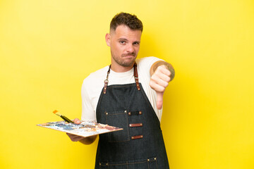 Young artist caucasian man holding a palette isolated on yellow background showing thumb down with negative expression