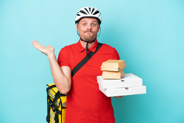 Young caucasian man with thermal backpack and holding fast food isolated on blue background having doubts while raising hands