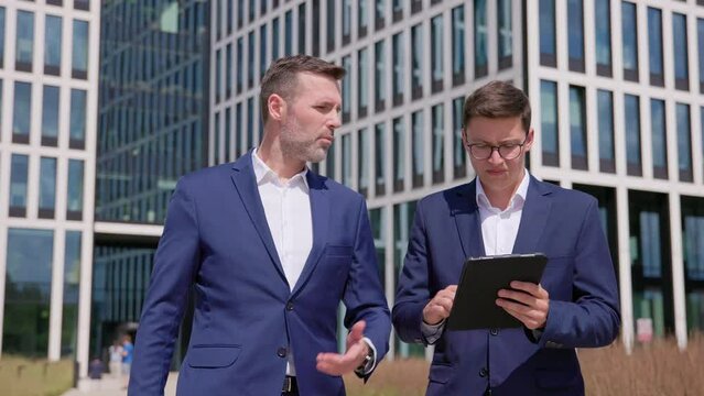Two men in blue suits use tablet, walk and talk outdoors, medium shot
