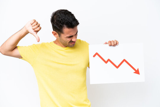 Young handsome man over isolated white background holding a sign with a decreasing statistics arrow symbol and doing bad signal