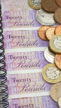 Vertical video social media format – Closeup of British sterling currency, next to a notebook and pen. £20 notes are laid out beneath £1 pound and other bronze and silver coins. Cash takings concept.