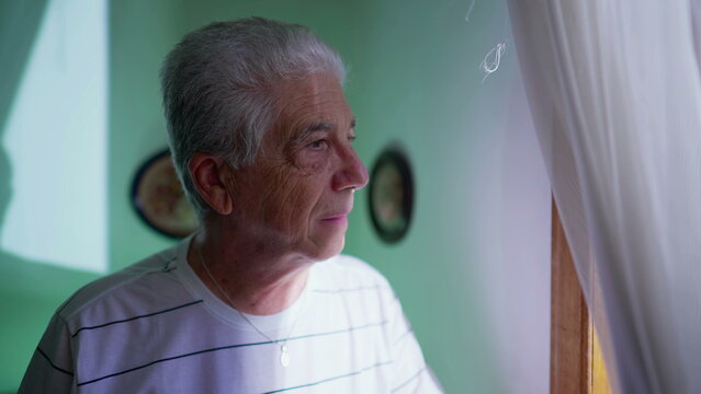 Senior Man Standing By Window At Home Looking Out View Through Curtains With Pensive Expression. Contemplative Elderly Gray-hair Man Watching Neighborhood From Residence