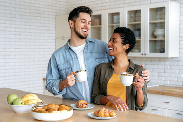 Fototapeta na wymiar Morning coffee. Happy couple in love in the morning. Multiracial spouses have tasty weekend breakfast in the kitchen at home with croissants and coffee, spend time together, communicate, smile