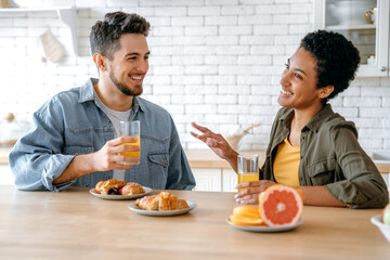 Fototapeta na wymiar Positive happy couple, an african american woman and a caucasian man, chat while sitting at home in the kitchen, smile, drink juice with croissants, spend leisure time together