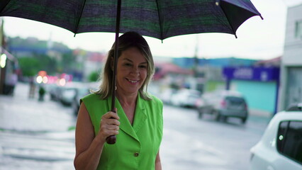 Happy middle-aged woman holding umbrella while walking in the rain city street sidewalk
