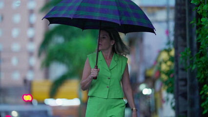 Pensive woman walking in the rain holding umbrella, strolling in urban environment sidewalk during bad weather. Person walks forward in street during rainy afternoon