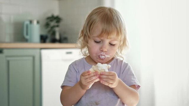 Baby girl enjoying ice cream. Pretty little toddler eating an ice-cream indoors, at home. Dining room background. Small child eats plombir and cream messy on her mouth. Cute kid with tasty sweet food.