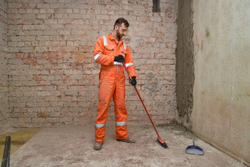 Young builder cleaning room from dust after demolition jobs, wearing gloves, boots and orange...