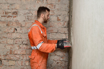 Man builder using h shape rule for plastering brick wall indoors, wearing gloves and orange coveralls.