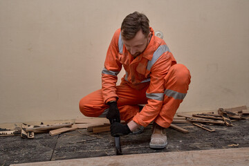 Young builder demolition old wooden parquet with crowbar, wearing gloves, boots and orange...