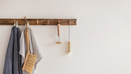 Wood hook rack with tea towels, broom and dish bottle brush hanging on white wall on the modern style kitchen background. Smart organisation storage ideas. Eco frienly banner. Natural material