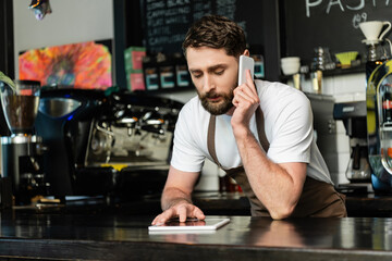 barista in apron talking on smartphone and using digital tablet on bar in coffee shop