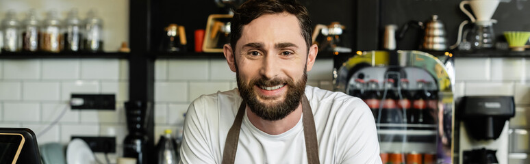 Obraz premium portrait of positive and bearded barista in apron looking at camera in coffee shop, banner