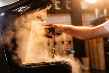 cropped view of barista in apron using coffee machine near steam and lighting in coffee shop