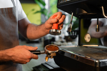 cropped view of barista in apron holding press and coffee in holder near coffee machine in cafe