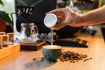cropped view of barista pouring sugar in cup of cappuccino near coffee beans in light in coffee shop