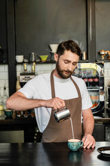 bearded barista in apron pouring milk from pitcher while making cappuccino on bar in coffee shop
