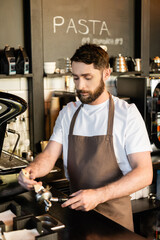 bearded barista in apron cleaning holder with brush near coffee machine while working in cafe