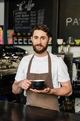 smiling bearded barista in apron holding cup of coffee and looking at camera in coffee shop