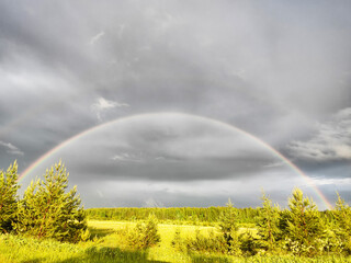 A rainbow over a green field and the sky in gloomy gray clouds on a spring, autumn or summer day or evening. The concept that the good and the bad things are together in the life