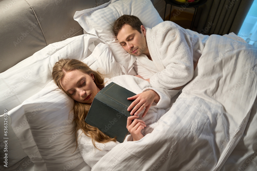 Wall mural Wife falling asleep reading book lying nearby sleeping husband