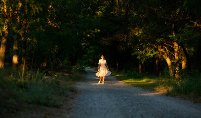 beautiful girl walking in forest, young woman in a dress stands on the path in sunlight among...