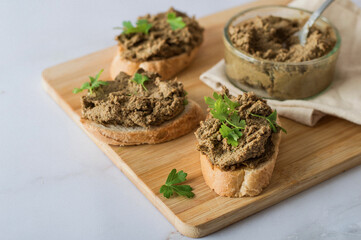 Liver pate spread on sliced ​​baguette, garnished with green onions, parsley and pretzel on a wooden board. Serving breakfast on a light background, close up.