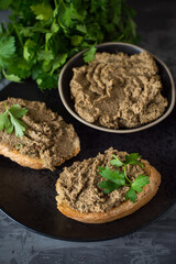Liver pate spread on a sliced ​​baguette, studded with parsley, a bowl with pate next to it. Filing against a dark background, close.